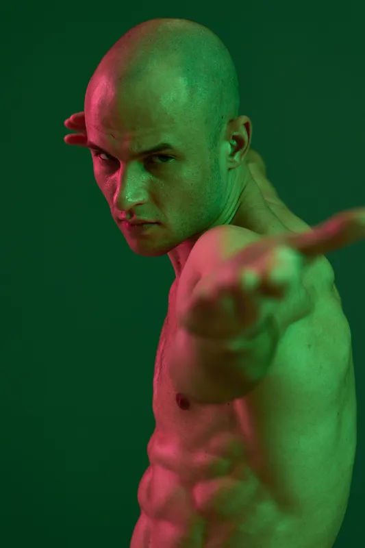 Dynamic man posing with intensity against a vibrant green background, showcasing his strong physique and emotional expression. Artistic lighting enhances the scene's impact.