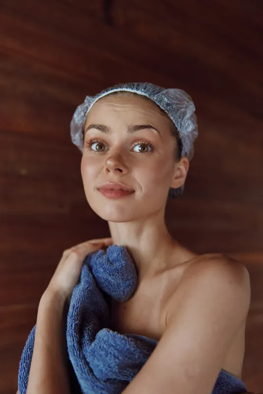 young woman with hair cap wrapped in blue towel inside wooden sauna, relaxed expression, natural lighting, warm tones, cozy atmosphere
