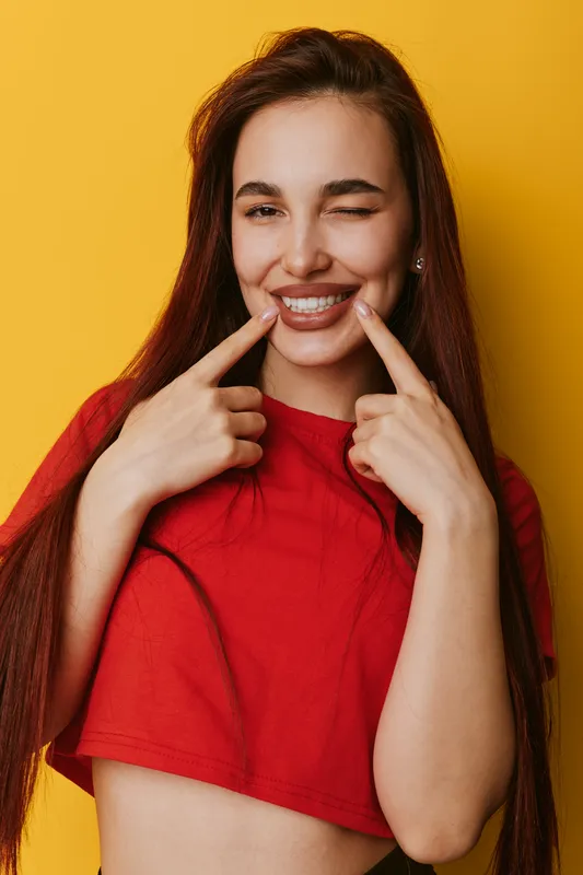 Young woman with long hair wearing red shirt smiling and winking while pointing fingers at her mouth on yellow background in studio portrait.