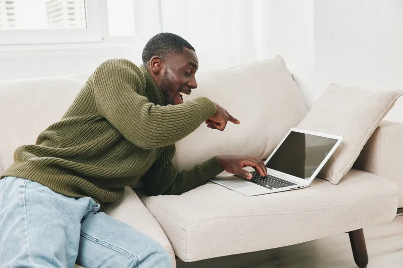 Cheerful young African man laughing while using a laptop on a cozy sofa in a bright living room, showcasing modern lifestyle and technology