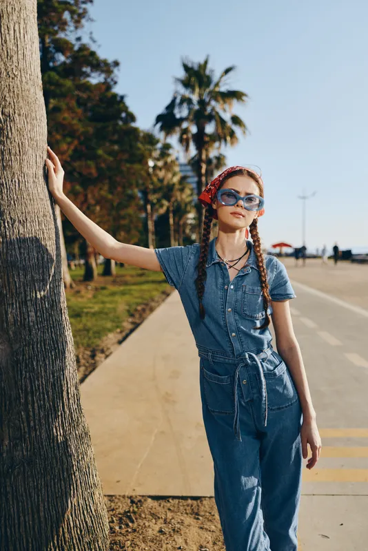 lifestyle woman in modern boho-western denim street style leans on a palm tree with sunglasses and red headband, film color tones enhancing casual summer vibe