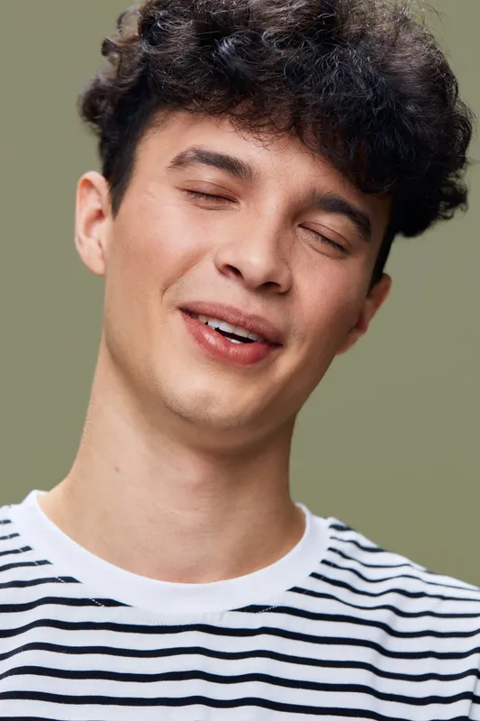 smiling man with curly hair wearing striped shirt, eyes closed, joyful expression, casual style, simple green background, natural light portrait