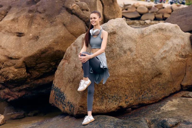 Young woman in activewear relaxing on a rocky beach with headphones, showcasing a healthy lifestyle and joyful mood, perfect for fitness and wellness themes