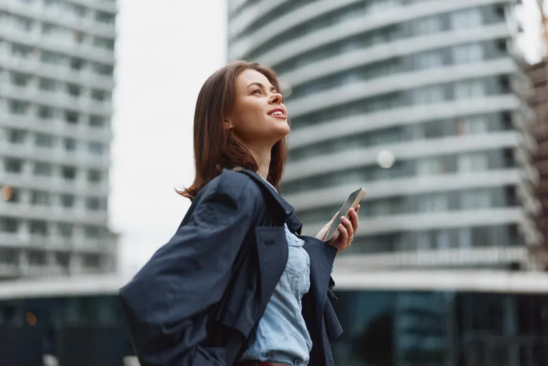 Cheerful woman outdoors, looking up with a smile, urban backdrop, daylight, casual professional outfit, breeze in hair, modern architecture, confident mood in urban setting.