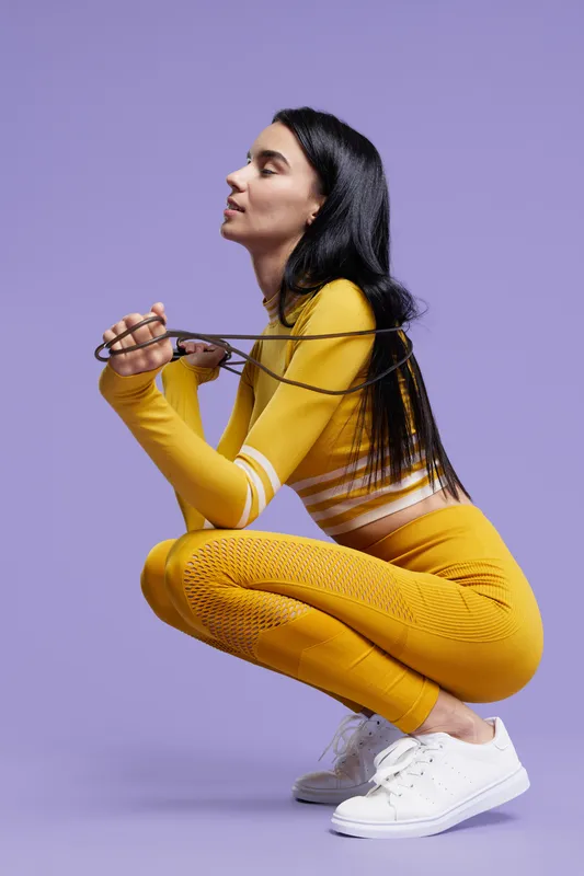 Woman sport fitness dressed in yellow activewear squatting while holding jump rope, focused and calm, on plain purple background, modern workout concept.