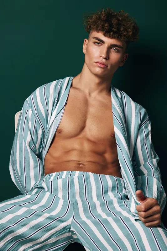 Young man with curly hair wearing striped open shirt and pants posing against dark green background with serious expression and toned body in studio.