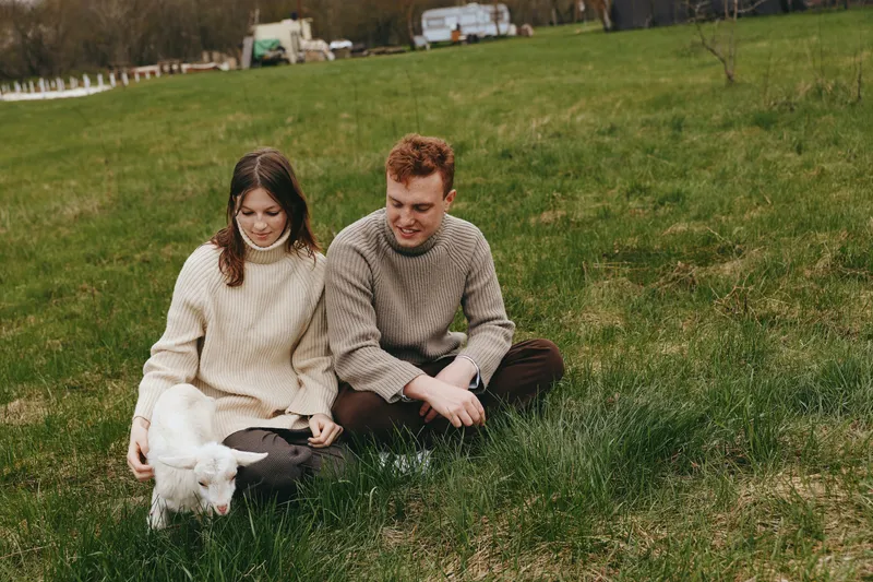 Cheerful couple outdoors, gentle goat kid nearby, warm sweaters, relaxed moment, soft sunlight, grassy meadow setting, candid interaction, simple natural colors, tranquil lifestyle scene