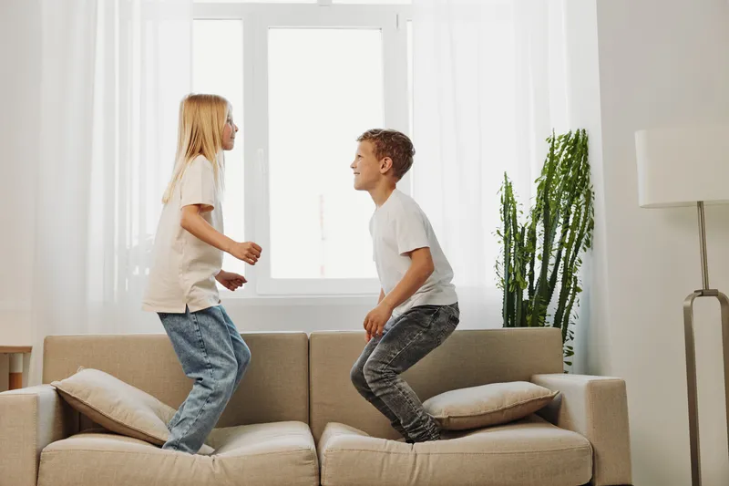 Two children jumping on a couch in a bright living room with natural light. The boy and girl enjoy playful moments together, capturing childhood energy and fun indoors.