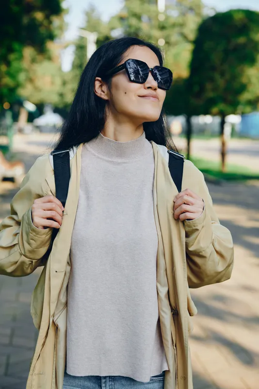 Woman wearing sunglasses and a light jacket stands outdoors in park, confident pose, street fashion style, casual chic look, sunlight highlights her contemporary wardrobe