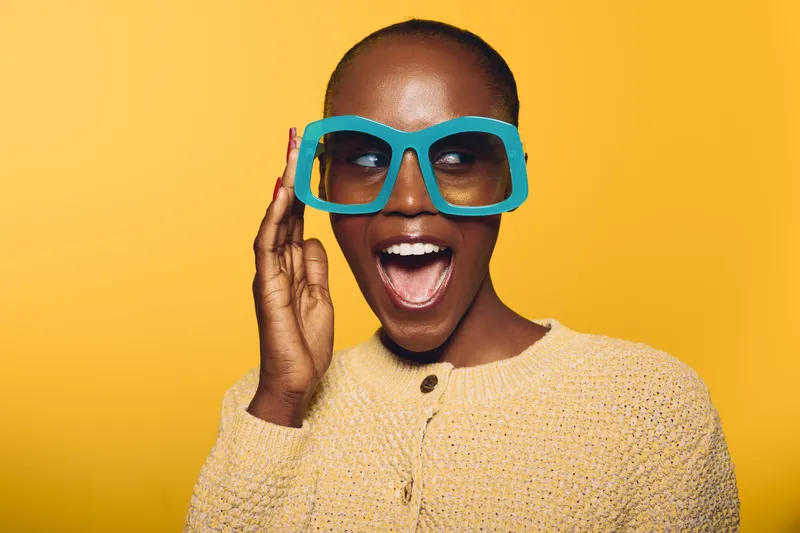 Excited dark skinned African woman wears oversized turquoise glasses and yellow sweater, posing with open mouth on yellow background. Concept of joy, fashion, and vibrant style.