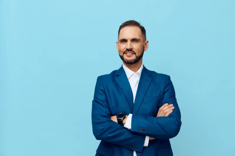 A confident business professional stands with arms crossed against a solid blue backdrop, exuding poise, focus, and readiness for leadership.