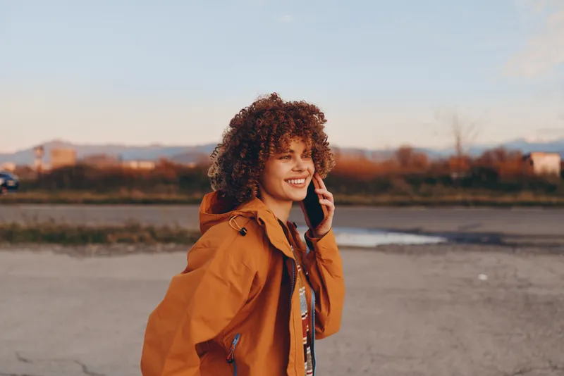 Woman lifestyle portrait outdoor at sunset, curly hair, orange jacket, smiling, speaking on phone near river path, warm evening light, calm mood, park background, travel