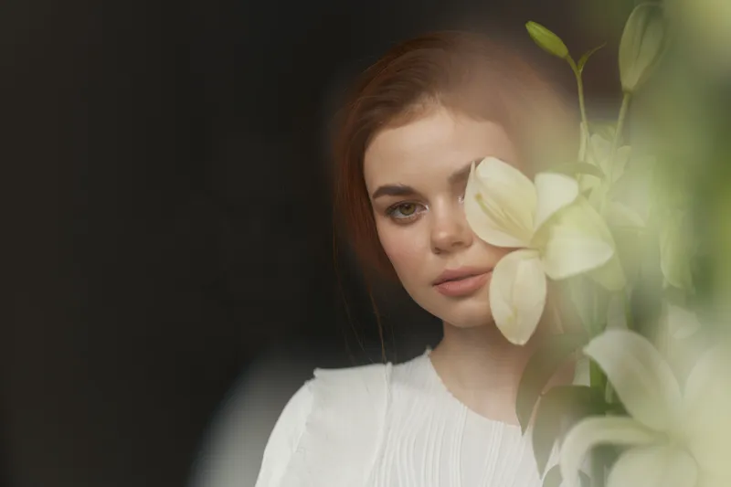 A young woman with light brown hair wearing a textured white blouse, softly peeking through white lilies, reflecting calm emotions and a serene vibe