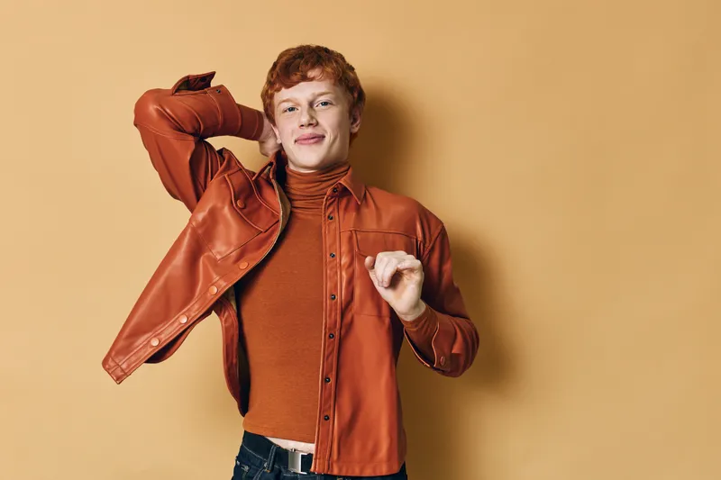 Young man with red hair wearing orange jacket and turtleneck poses confidently against beige background in studio portrait for fashion or lifestyle concept and style.