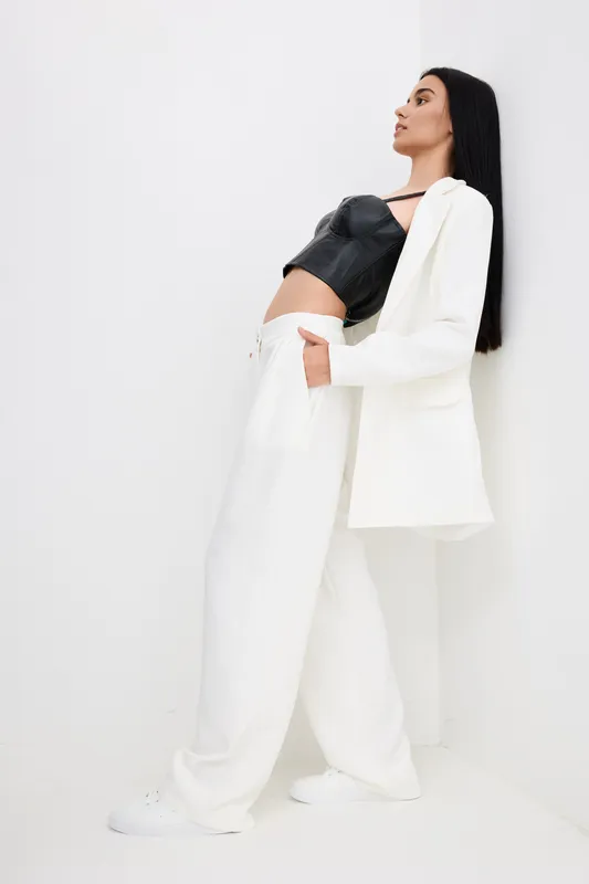 tall woman in black top and white suit poses confidently against white wall, showcasing modern fashion and minimalistic style with relaxed mood and clean background