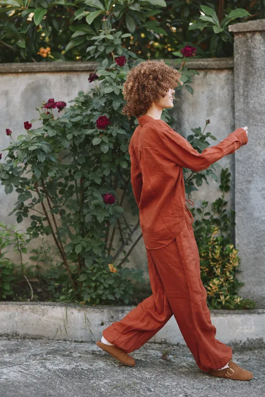 Woman in rust suit walking along garden wall, casual lifestyle portrait, natural light, soft greenery and roses in the background, candid street style