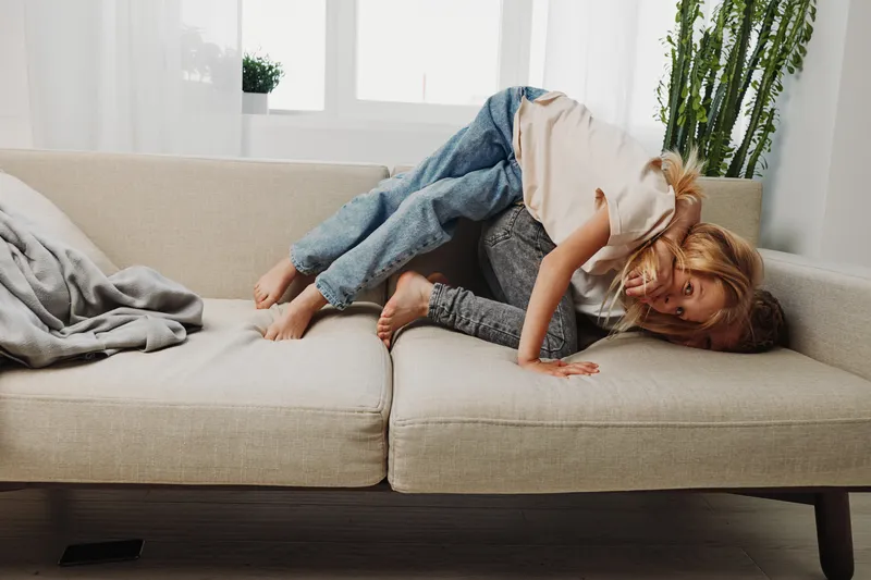 Playful children having fun on beige sofa in bright living room. Siblings enjoy active moment together, laughing and climbing over each other. Concept of family joy and childhood play.