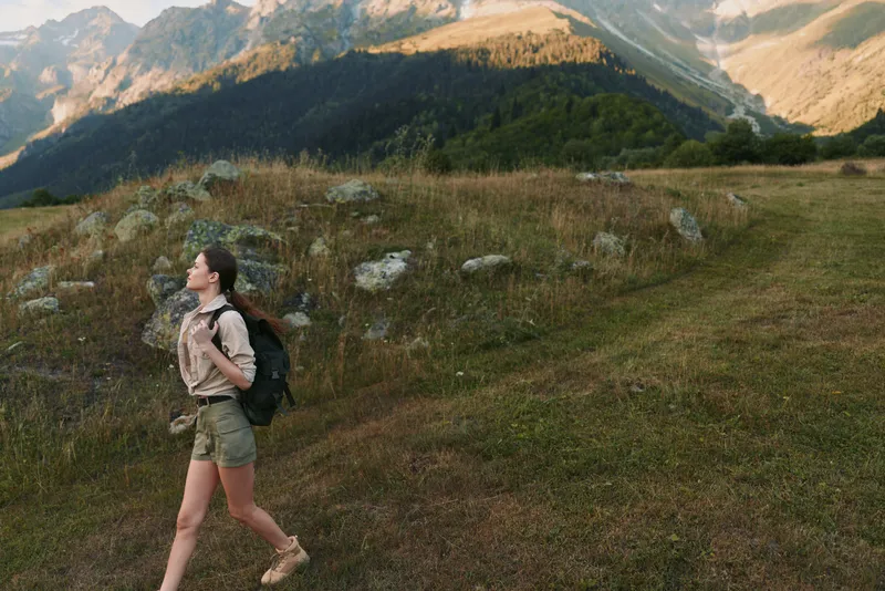 Young woman hiking in a mountainous meadow with a backpack, looking up at the peaks with a sense of discovery