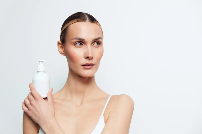 Young woman with clear skin holding white lotion bottle, minimal makeup, neutral background, skincare concept, clean beauty, fresh and natural look, soft lighting