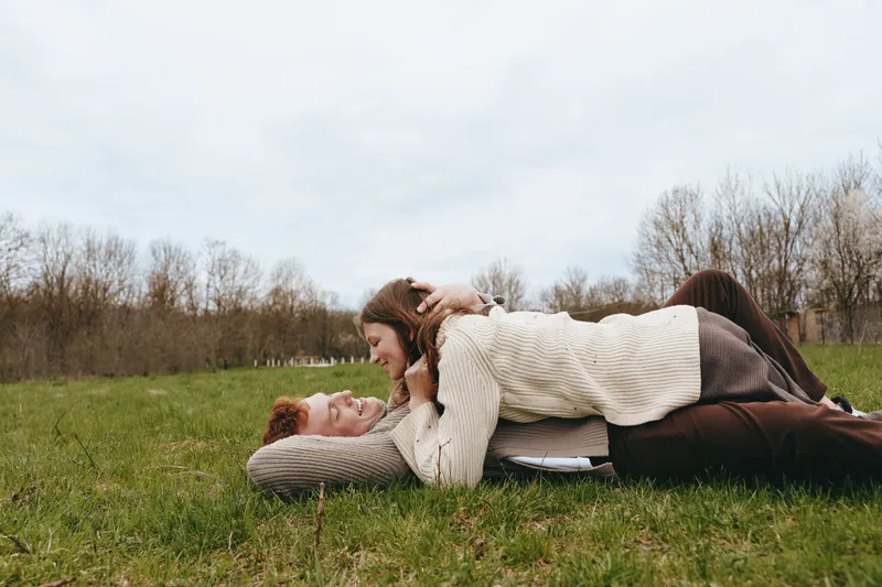 Couple outdoors on grass, playful embrace in a relaxed moment, cozy sweater and natural tones, candid affection under muted sky, park scenery and wide meadow, calm and warm mood, lifestyle moment