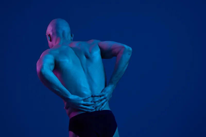 Athletic man with a muscular build experiencing discomfort, showcasing dramatic lighting with shades of blue and purple, highlighting the tension in his posture.