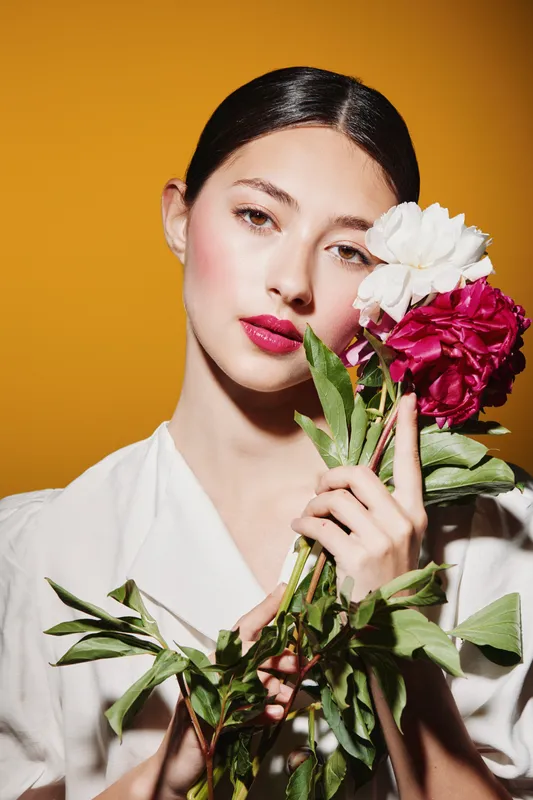 Young woman with bright pink lipstick and smooth skin holds delicate white and red flowers near her face. Studio shot with vibrant orange background focused on beauty and fashion.