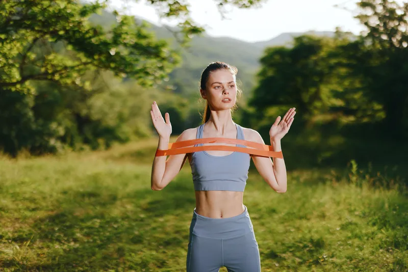 Athletic woman exercising outdoors in sportswear with resistance bands, showcasing strength and focus in a lush green natural setting