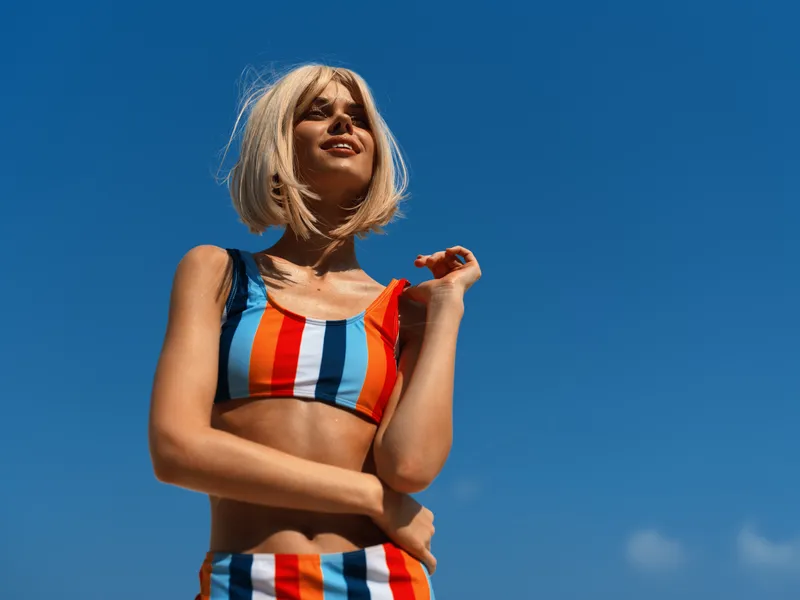 confident young woman wearing colorful striped swimsuit standing outdoors, enjoying sunny weather with clear blue sky, summer beach vibe, close-up portrait