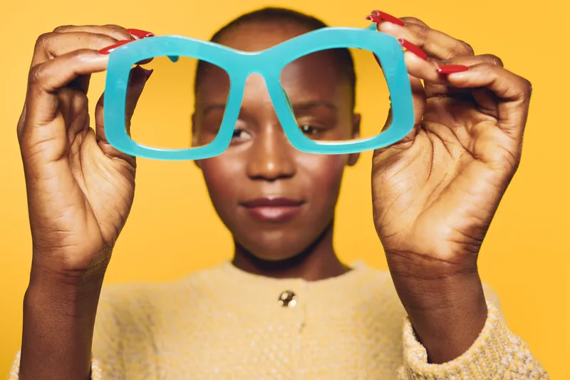 Young black woman with dark skin holds large blue eyeglasses in front of her face on yellow background. Concept of vision, fashion, and style with focus on accessories and portrait.