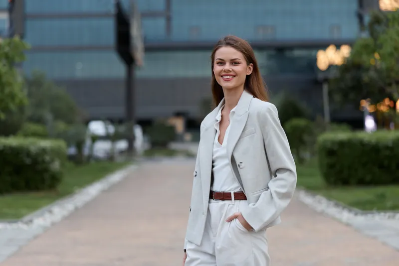 Confident young woman in light gray business suit smiling outdoors in urban park setting on warm day