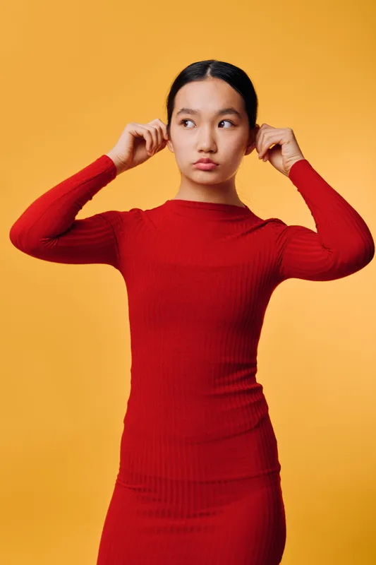 Woman in red dress adjusting white earphones with thoughtful expression standing against orange background. Fashion, technology, and casual style in modern portrait illustration.
