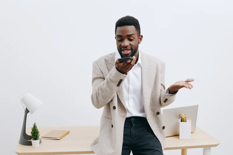 Professional young African American man in casual jacket and white shirt standing at desk using mobile phone with a laptop, pens, notebook, and minimal decor, inside a modern office space, casual busi