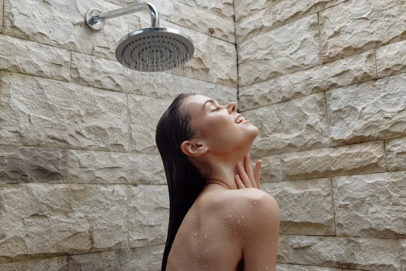 young woman enjoying outdoor shower under rainfall with closed eyes, wet hair, stone wall background, feeling relaxed and refreshed in natural light