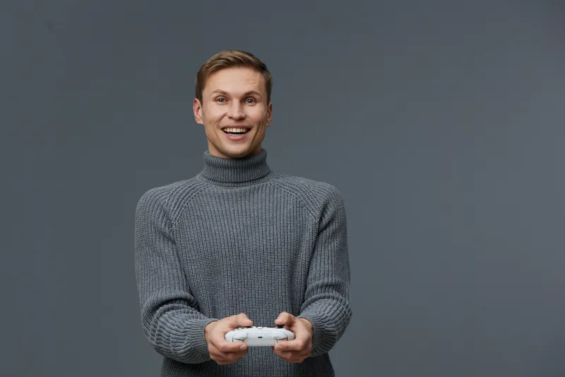 young smiling man hold game controller wear grey knitted turtleneck sweater isolated on grey background play digital entertainment technology concept
