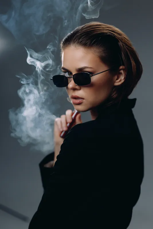 Stylish woman wearing sunglasses and smoking with rising smoke in a dark setting, conveying mystery and attitude.