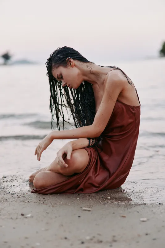 A graceful woman sits on a sandy beach at dusk, with wet hair and a flowing dress, capturing a moment of calm reflection and subtle vulnerability by the shore.
