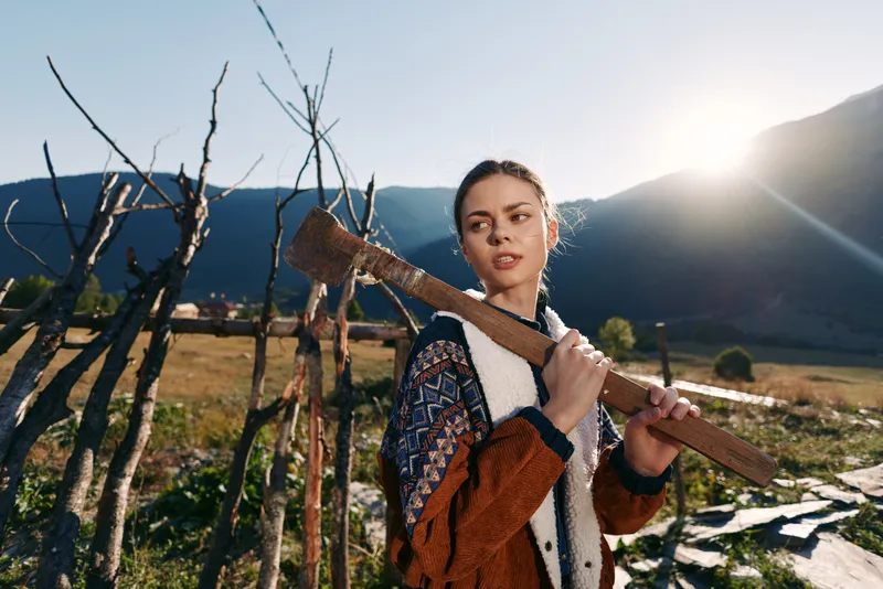 Woman carrying an axe over her shoulder in rural countryside with mountains and bright sunlight, farming fence and fields, outdoor portrait of a young worker in nature.