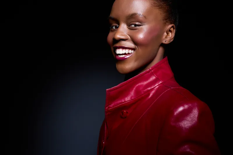 Smiling young black woman with dark skin wearing a red leather jacket poses against a black background. Concept of beauty, fashion, confidence, and stylish portrait.