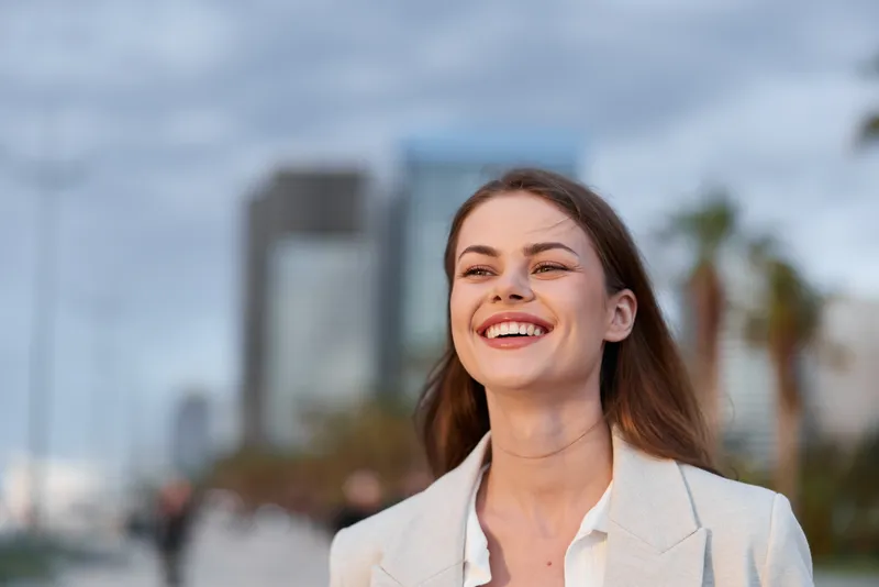 Confident businesswoman smiling in a cityscape, wearing a light blazer, with a warm sunset glow, conveying professionalism and positivity.