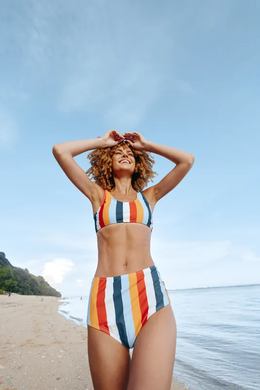 Woman beach bikini ocean summer sunlight curly hair hands on head relaxing outdoors, standing on sandy shoreline facing sky, smiling in striped swimwear, calm sea background, horizon