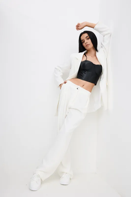 stylish woman posing confidently in black corset and white suit against minimalist white background, showcasing modern fashion and attitude