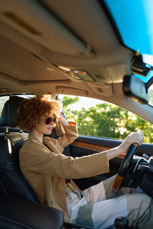 Woman driver in a stylish car, daytime light, relaxed lifestyle moment, sunglasses on, curly hair, beige jacket, open road in view, candid and bright atmosphere