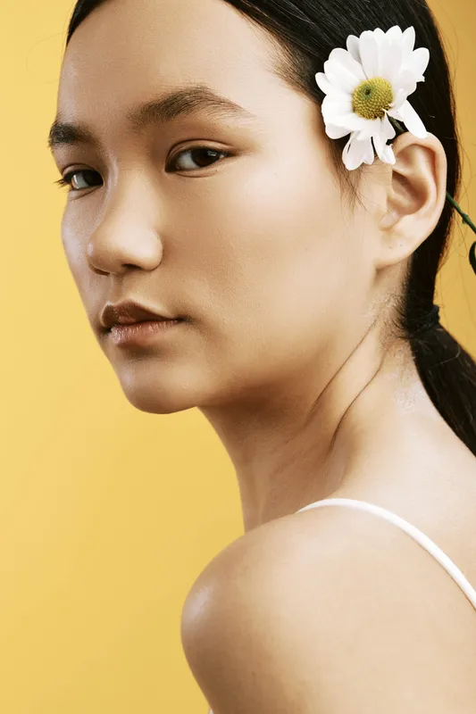 Asian woman is captured in a closeup portrait against a warm yellow backdrop, with a delicate flower tucked behind the ear. The serene expression and soft lighting emphasize natural beauty and calm