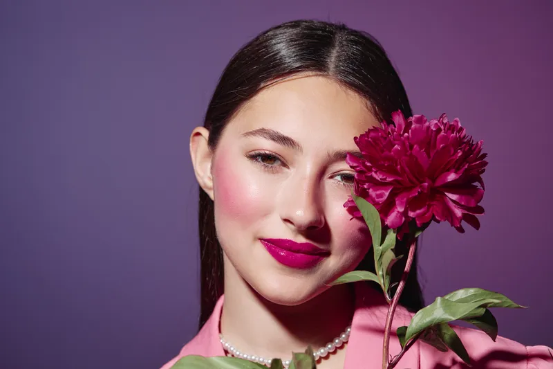 Young woman with glossy dark hair poses with a lush pink flower cover on violet background. Artful makeup with vivid lipstick and soft pearl necklace, concept of beauty fashion creative.