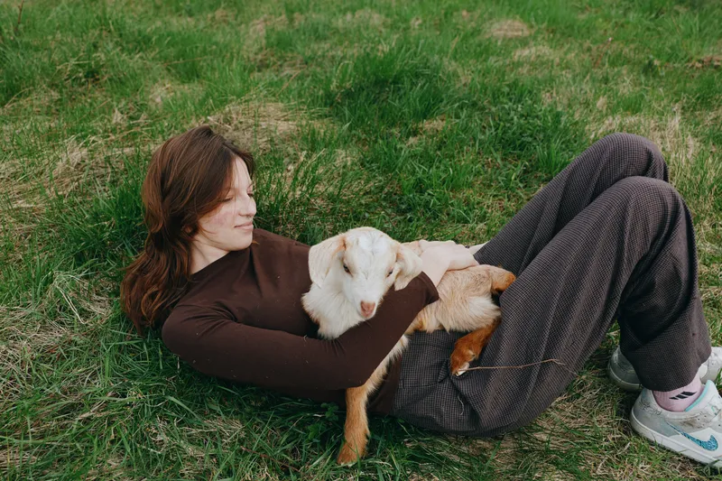 Relaxed woman lying on grass with a goat kid resting on her lap in outdoors, a calm countryside portrait of nature and relaxation, showing cuddle, bond, and warm happiness in a sunny, peaceful