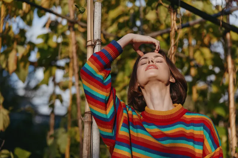 Woman in a rainbow sweater leaning against a wooden post outdoors, short hair, mid-20s, relaxed and blissful with closed eyes in warm sunlight, casual fashion lifestyle.