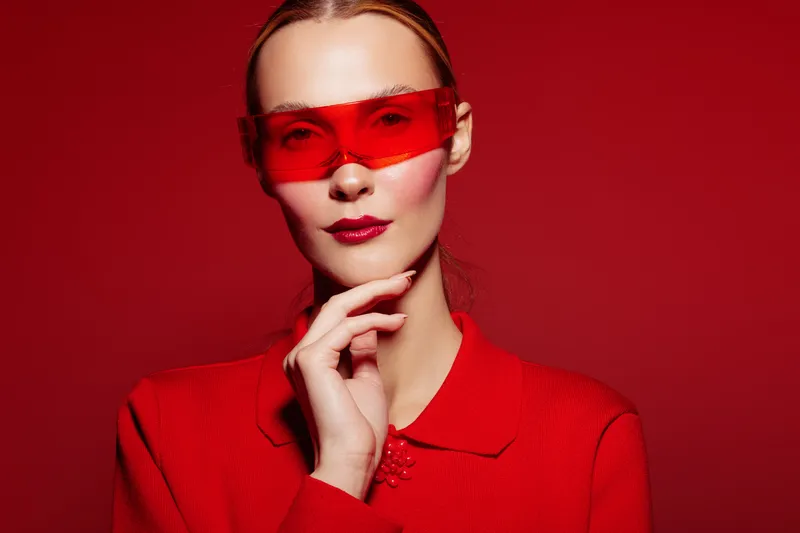 Stylish woman in red sweater and red translucent glasses poses against red background. Fashion portrait with bold color contrast and modern accessories. Concept of style and confidence.