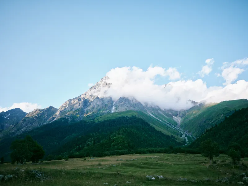 Mountains rise over a grassy meadow beneath a clear blue sky with fluffy clouds, creating a tranquil alpine landscape ideal for nature, travel, and outdoor inspiration in stock photography