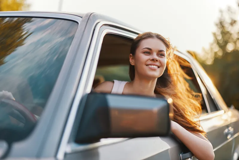 Young woman enjoying a drive with her head out of the car window, conveying joy and freedom in a sunny setting, with flowing hair and a vibrant nature backdrop.