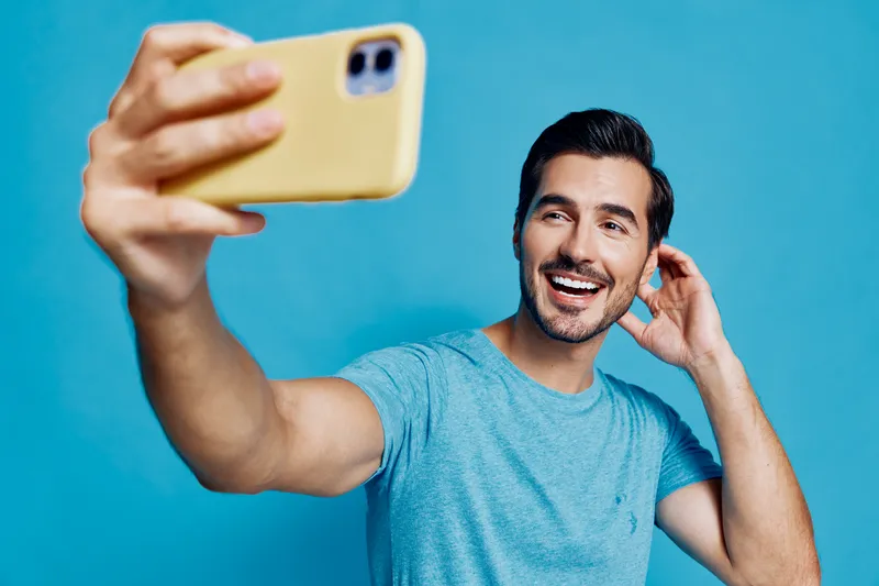 Happy smiling young man taking a selfie with smartphone in hand against vibrant blue background, wearing casual t-shirt. Lifestyle concept.